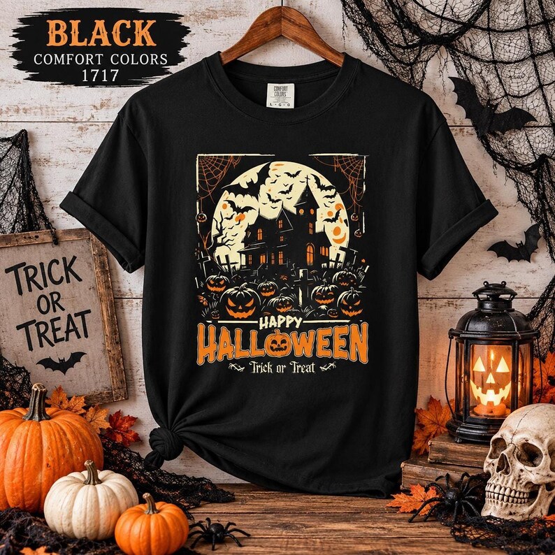 A black t-shirt laid flat on a wooden surface with a haunted house and pumpkin graphic, surrounded by Halloween props including pumpkins and a skull.