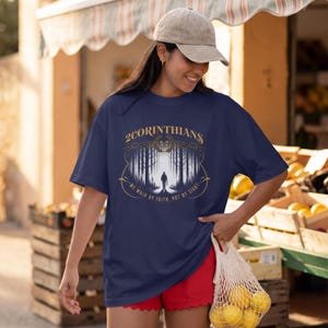 Könnte beinhalten: Ein marineblaues T-Shirt mit einer Grafik einer Person, die durch einen Wald geht, sowie dem Text 2CORINTHIANS 5:7 und WE WALK BY FAITH, NOT BY SIGHT. Das Design enthält einen Lorbeerkranz und dekorative goldene und weiße Verzierungen.