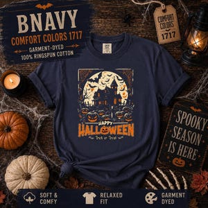 A navy blue t-shirt laid flat on wood with a haunted house and pumpkin graphic, styled with miniature pumpkins and a lantern.