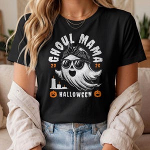 May include: A black short-sleeve t-shirt featuring a white graphic of a ghost wearing sunglasses and a bandana. The text reads GHOUL MAMA, 20 26, and HALLOWEEN, accompanied by illustrations of three candles and two orange jack-o'-lanterns.