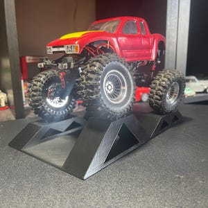 May include: A red and yellow toy monster truck with large, knobby tires is displayed on a black ramp. The truck has a detailed body and sits on a black surface. The tires are large and rugged.