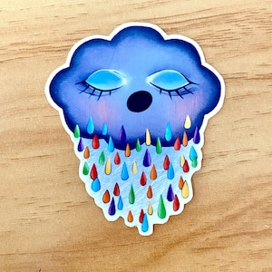 May include: A sticker featuring a cartoon cloud with closed eyes and colorful raindrops. The cloud is a gradient of blue hues, with a black mouth and blue eyes. The raindrops are various colors, including red, orange, yellow, green, and blue.
