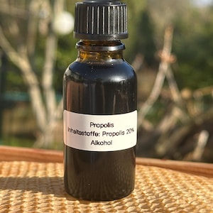 May include: A small, dark amber glass bottle with a black cap. A white label displays the word "Propolis" and ingredient information. The bottle is filled with a dark liquid, likely a health supplement or extract.