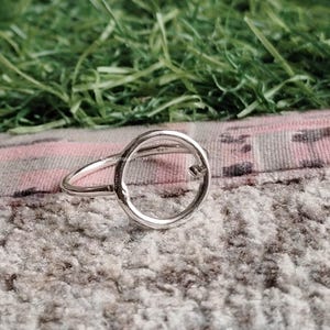 Puede incluir: Un anillo de plata con un diseño circular. El anillo presenta una banda fina y un círculo abierto prominente en el centro. El anillo se muestra sobre una superficie texturizada con tonos verdes y rosas en el fondo.