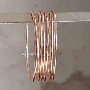 Może przedstawiać: Kolekcja cienkich, różowo-złotych bransoletek. Bransoletki są ułożone w stos, prezentując swój delikatny design i ciepły metaliczny odcień. Biżuteria jest prezentowana na srebrnym pręcie. Widoczna jest nazwa marki "ROYALARTISANJEWELRY".
