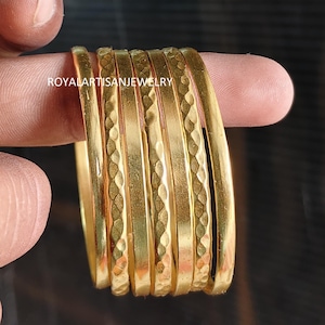 May include: A set of six gold-colored bangles featuring a hammered texture on the outer surface. The bangles are arranged in a stack, and the text ROYALARTISANJEWELRY is visible on the surface of the metal. The bangles have a polished finish.