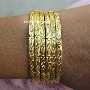 Puede incluir: Un conjunto de seis pulseras doradas con diseños florales y de hojas. Las pulseras se llevan en la muñeca y tienen una superficie brillante y reflectante. Las joyas son probablemente de metal y son un accesorio decorativo.