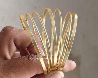Könnte beinhalten: Ein Stapel goldfarbener Armreifen. Die Armreifen bestehen aus dünnem, flachem Metall und sind in einer gebogenen, fächerartigen Form angeordnet. Der Schmuck wird in der Hand einer Person gehalten. Der Markenname "ROYALARTISANJEWELR" ist sichtbar.