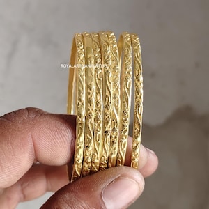 Peut inclure: Un ensemble de six bracelets dorés ornés de motifs floraux et de vignes complexes. Les bracelets sont tenus dans une main, mettant en valeur leur design détaillé et leur finition polie. Les bijoux sont présentés sur un fond neutre.