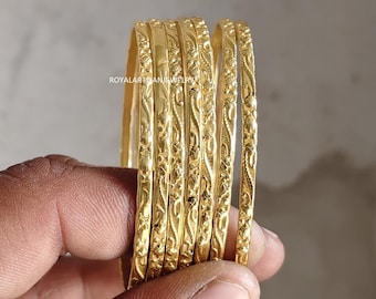 Könnte beinhalten: Ein Satz von sechs goldenen Armreifen mit aufwendigen Blumen- und Rankenmustern. Die Armreifen werden in einer Hand gehalten, wodurch ihr detailliertes Design und ihre polierte Oberfläche hervorgehoben werden. Der Schmuck wird vor einem neutralen Hintergrund präsentiert.