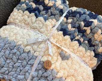 Puede incluir: Una manta tejida a mano, gruesa, en tonos azules y crema, atada con una cinta blanca. La manta tiene un aspecto suave y texturizado y una pequeña etiqueta que dice "Hecho a mano con amor".
