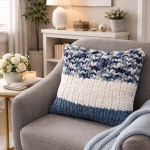 May include: A square, chunky-knit pillow in shades of blue and white rests on a gray armchair. The pillow features a striped design with a dark blue base, a white center, and a patterned top section in varying shades of blue.