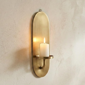 May include: A wall-mounted candle holder with a brass finish. The holder has an oval shape with a small circular base to hold a white candle. The candle is lit, casting a warm glow. The holder is attached to the wall with a small loop.