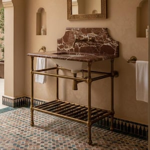 May include: A bathroom with a vintage design. A red marble sink sits on a brass frame, with a white basin. A decorative mirror hangs above the sink, and two sconces flank it. The walls are a warm beige, and the floor has a patterned tile.