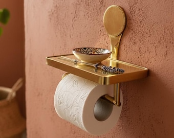 Könnte beinhalten: Ein goldfarbener Toilettenpapierhalter aus Metall mit einem kleinen Regal, das eine Rolle weißes Toilettenpapier hält. Eine kleine dekorative Schale und ein Lavendelzweig liegen auf dem Regal. Der Halter ist an einer strukturierten, rotbraunen Wand befestigt.