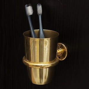 May include: A gold-colored toothbrush holder with two gray toothbrushes. The holder is cylindrical with a rounded base and a mounting bracket. The toothbrushes have white bristles.