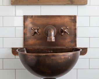 May include: A copper-toned wall-mounted sink with a matching faucet and cross-shaped handles. The sink has a rounded basin and is set against a white tiled wall. A wooden framed mirror hangs above the sink.