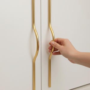 May include: Gold-colored cabinet pulls with a wavy design. The pulls are mounted on a white cabinet door. The image shows two pulls, one of which is being held by a hand.