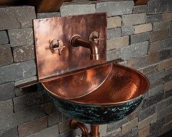 May include: A copper wall-mounted sink with a hammered finish. The sink has a curved basin with a dark green patina around the rim. The faucet and fixtures are also copper, mounted on a rectangular copper backplate. The sink is installed on a stone wall.