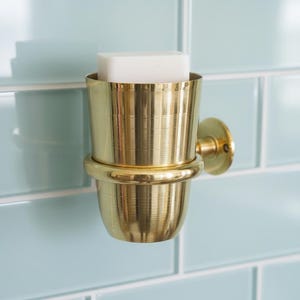 May include: A gold-colored, wall-mounted soap dish with a cylindrical design. The dish holds a white bar of soap and is set against a light blue tiled wall. The fixture has a classic, elegant appearance.
