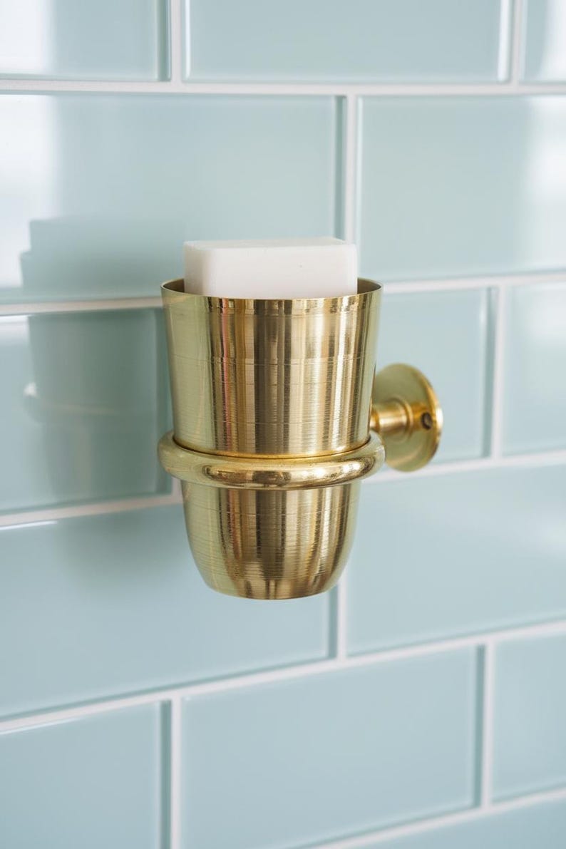 May include: A gold-colored, wall-mounted soap dish with a cylindrical design. The dish holds a white bar of soap and is set against a light blue tiled wall. The fixture has a classic, elegant appearance.