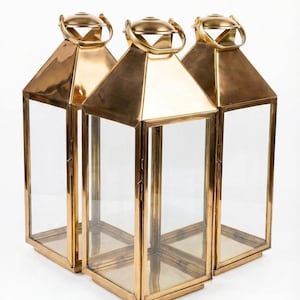 May include: Three gold-colored metal lanterns with clear glass panels. Each lantern has a geometric shape with a handle at the top. The lanterns are arranged on a white background.