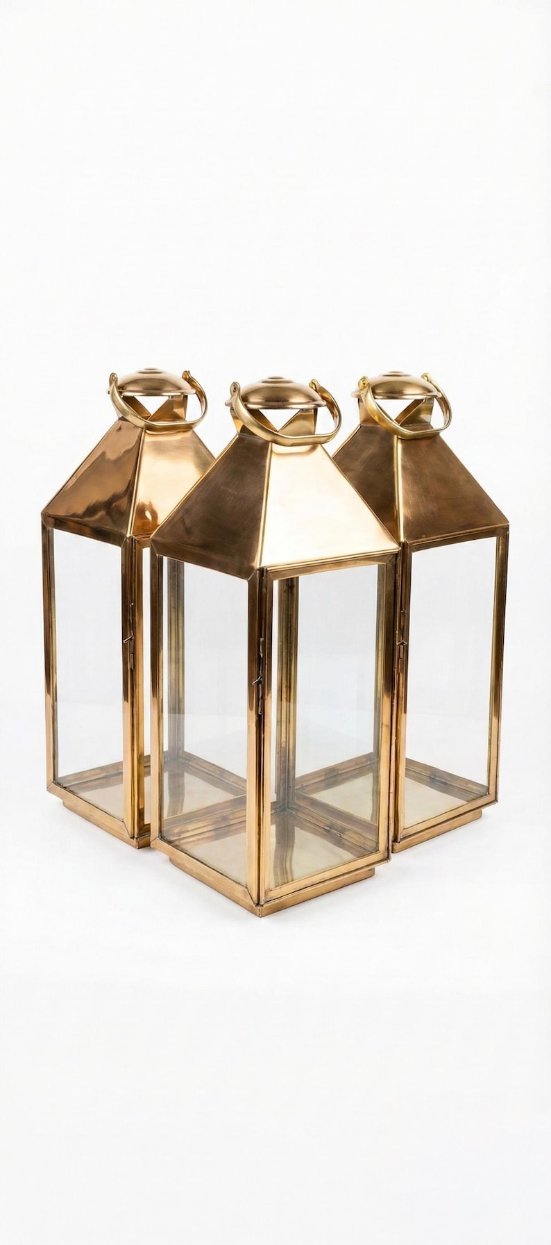 May include: Three gold-colored metal lanterns with clear glass panels. Each lantern has a geometric shape with a handle at the top. The lanterns are arranged on a white background.