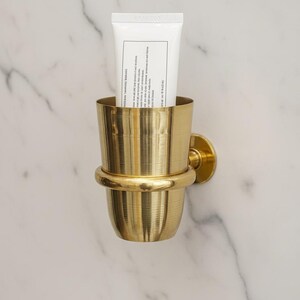 May include: A gold-colored metal toothbrush holder mounted on a marble wall, holding a white tube. The holder has a cylindrical cup shape with a decorative band and a circular wall mount.