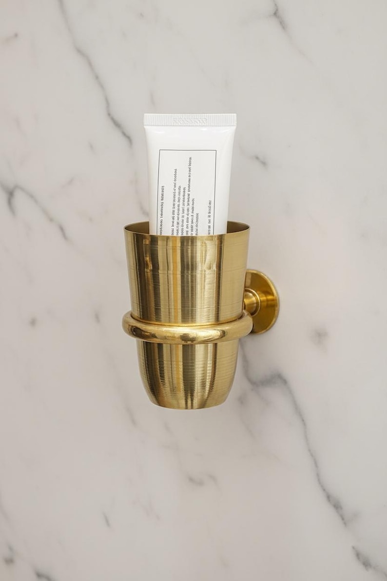 May include: A gold-colored metal toothbrush holder mounted on a marble wall, holding a white tube. The holder has a cylindrical cup shape with a decorative band and a circular wall mount.