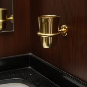May include: A gold-colored metal toothbrush holder mounted on a dark wood wall. The holder has a cylindrical cup shape with a ring around the middle and a circular base. The bathroom fixture is near a black countertop.