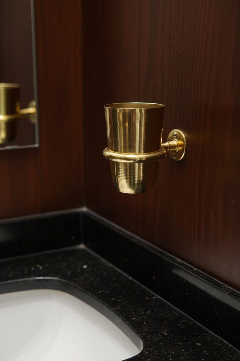 May include: A gold-colored metal toothbrush holder mounted on a dark wood wall. The holder has a cylindrical cup shape with a ring around the middle and a circular base. The bathroom fixture is near a black countertop.