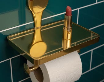 Könnte beinhalten: Ein goldfarbenes Badezimmeraccessoire mit Ablage, Toilettenpapierhalter und Lippenstift. Das Accessoire ist an einer türkisfarbenen gefliesten Wand angebracht. Der Lippenstift hat einen gedämpften Rosaton.