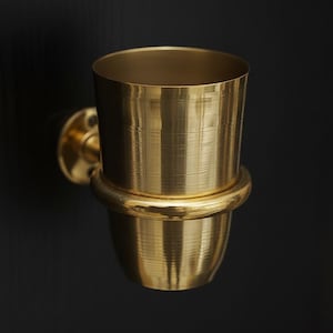 May include: A gold-colored, wall-mounted toothbrush holder. The holder has a cylindrical cup shape with a rounded base and a circular mounting plate. The metallic surface reflects light, creating a warm, inviting aesthetic. The design is simple and elegant.