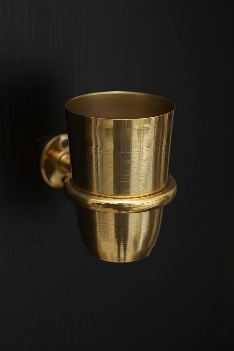 May include: A gold-colored, wall-mounted toothbrush holder. The holder has a cylindrical cup shape with a rounded base and a circular mounting plate. The metallic surface reflects light, creating a warm, inviting aesthetic. The design is simple and elegant.