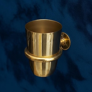 May include: A gold-colored, wall-mounted toothbrush holder. The holder has a cylindrical cup shape with a rounded base and a circular mounting plate. The holder is made of metal and has a shiny finish. The background is a dark blue.