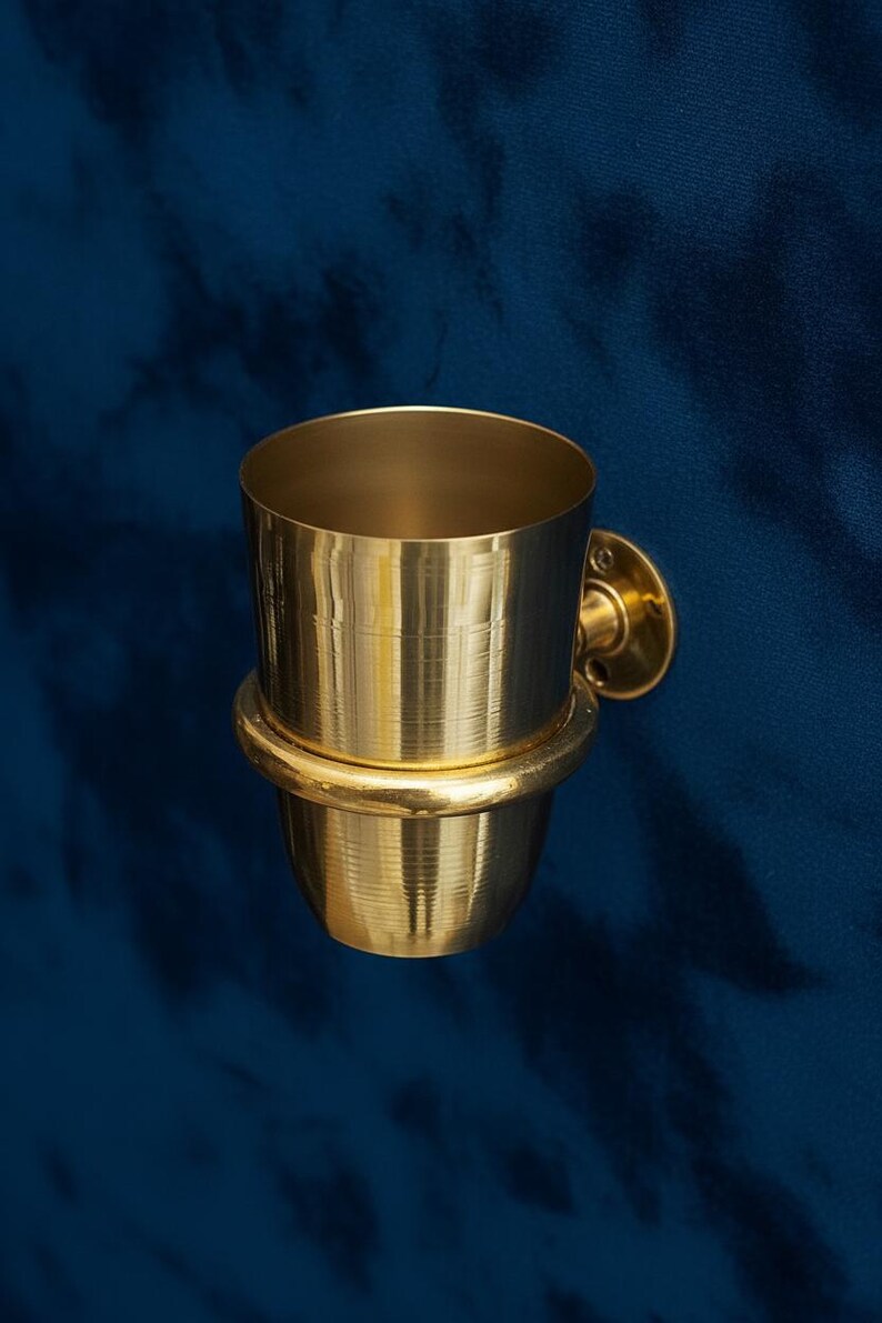 May include: A gold-colored, wall-mounted toothbrush holder. The holder has a cylindrical cup shape with a rounded base and a circular mounting plate. The holder is made of metal and has a shiny finish. The background is a dark blue.
