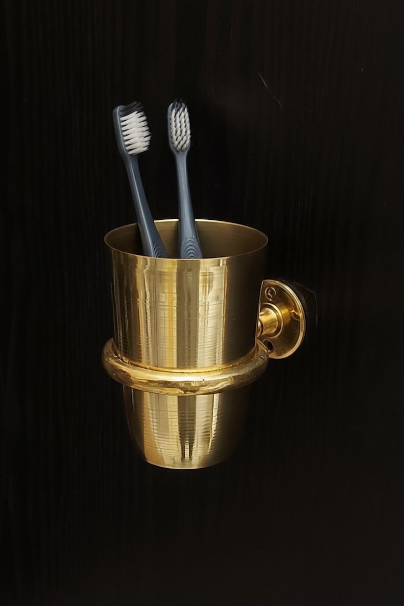 May include: A gold-colored toothbrush holder with two gray toothbrushes. The holder is cylindrical with a rounded base and a mounting bracket. The toothbrushes have white bristles.