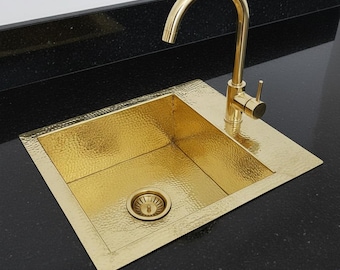 May include: A hammered gold kitchen sink with a matching faucet. The sink is rectangular with a textured surface and a drain. The faucet has a curved spout and a single lever handle. The sink is set into a black countertop.