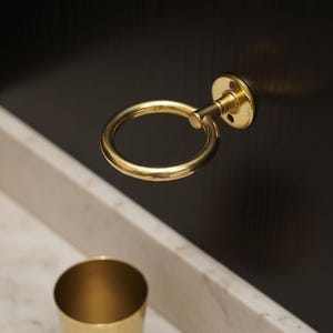 May include: A close-up of a gold-colored, circular cabinet pull with a matching cup. The pull is mounted on a dark wooden surface. The cup is sitting on a white marble surface.