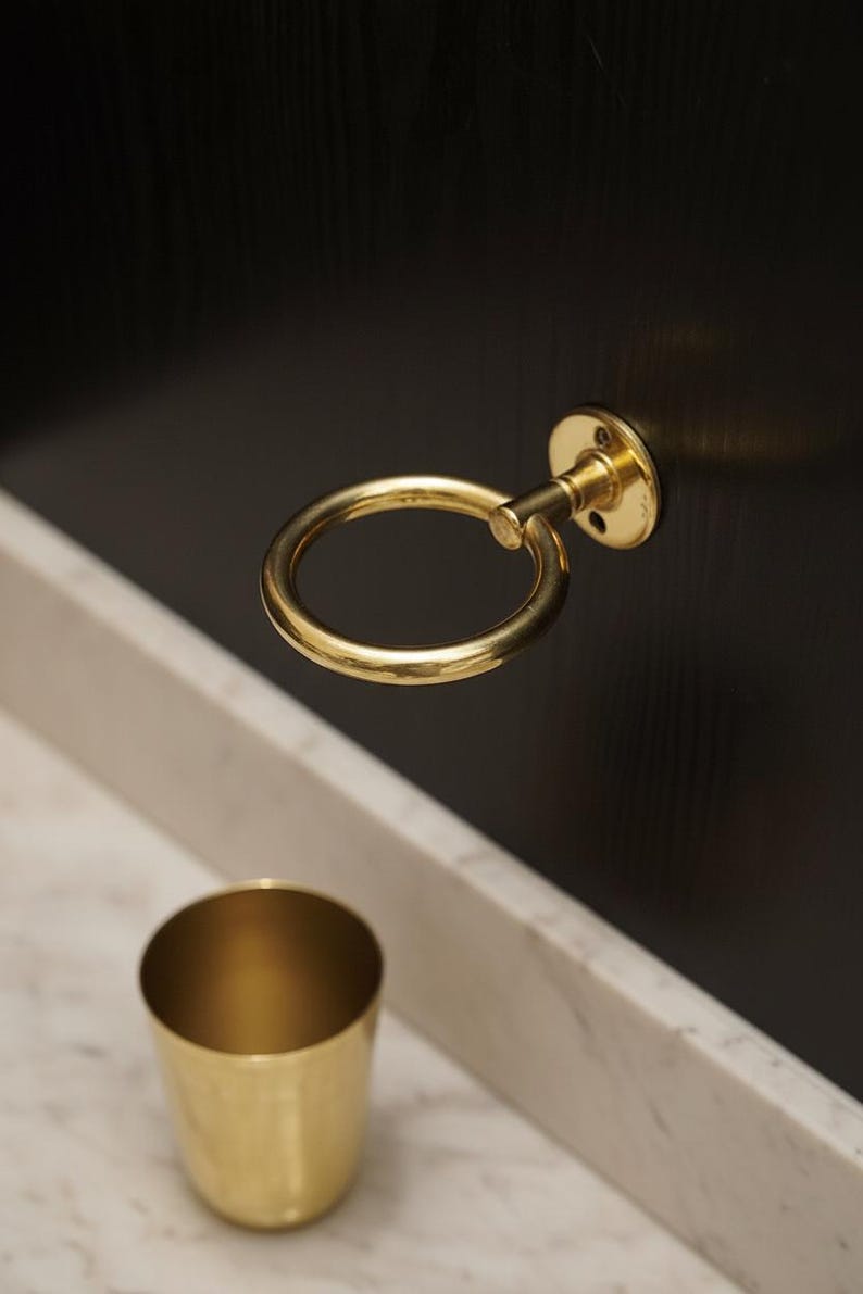 May include: A close-up of a gold-colored, circular cabinet pull with a matching cup. The pull is mounted on a dark wooden surface. The cup is sitting on a white marble surface.