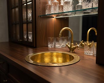 May include: A hammered gold sink basin with a matching faucet is set into a dark wood countertop. Crystal glassware and decanters are displayed on shelves in the background, creating a luxurious bar area.