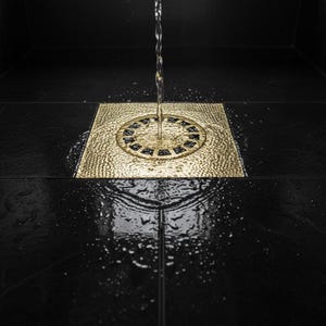 Peut inclure: Gros plan d'une bonde de douche carrée dorée avec de l'eau qui s'y déverse. La bonde est encastrée dans un carrelage noir. Des gouttelettes d'eau sont visibles sur la surface du carrelage, reflétant la lumière.