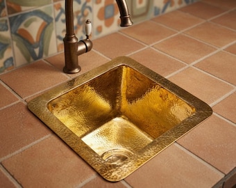 May include: A hammered gold-colored square sink with a bronze faucet. The sink is set into a countertop of reddish-brown tiles. The faucet has a curved spout and a white handle. The background features decorative tiles in shades of blue, orange, and brown.