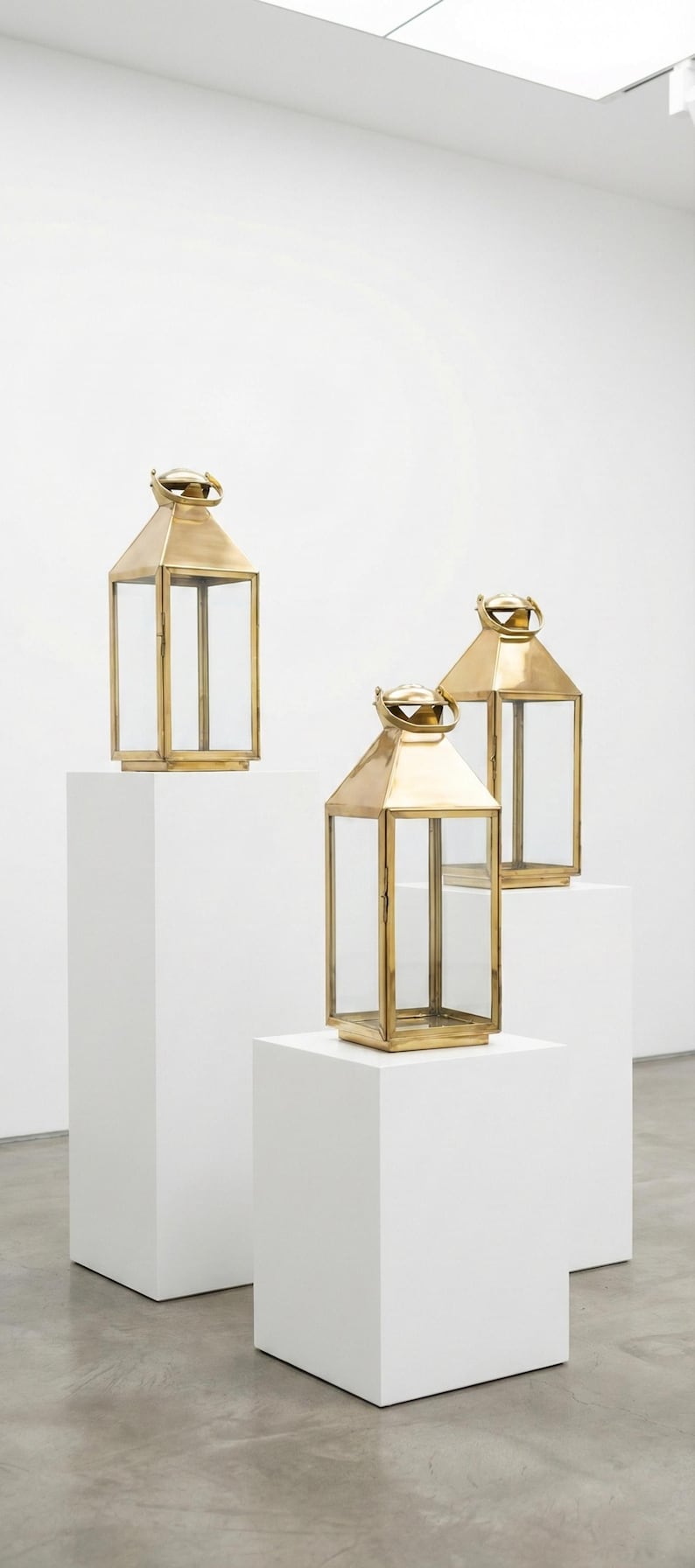 May include: Three gold-colored lanterns with clear glass panels are displayed on white pedestals. Each lantern has a rectangular shape with a peaked roof and a handle at the top. The lanterns are in a well-lit, minimalist setting.