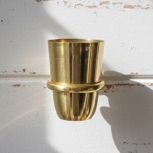 May include: A gold-colored, two-part metal container. The top section is cylindrical, and the bottom section is a rounded cup shape. The two parts are connected by a thick, rounded band. The container is set against a white, textured background.