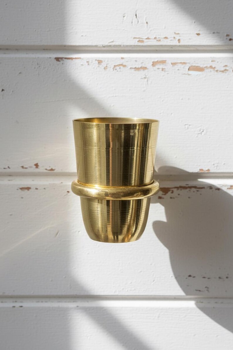 May include: A gold-colored, two-part metal container. The top section is cylindrical, and the bottom section is a rounded cup shape. The two parts are connected by a thick, rounded band. The container is set against a white, textured background.