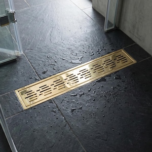 May include: A rectangular, gold-colored shower drain with a slotted design is installed on a dark gray tiled floor. Water droplets are visible on the tiles, suggesting recent use. The drain is positioned near a glass shower enclosure.