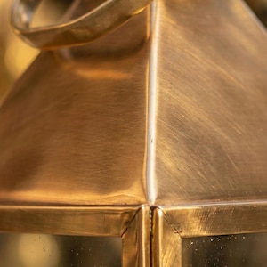 May include: Close-up of a vintage brass lantern with a handle. The lantern has a rectangular shape with a peaked top and clear glass panels. The brass has a warm, golden hue.