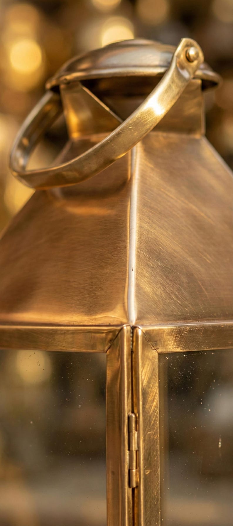May include: Close-up of a vintage brass lantern with a handle. The lantern has a rectangular shape with a peaked top and clear glass panels. The brass has a warm, golden hue.