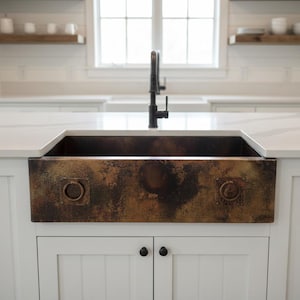 May include: A copper farmhouse sink with a dark patina finish. The sink has a rectangular basin and is mounted on a white cabinet with black hardware. The kitchen features a white countertop and a black tap.